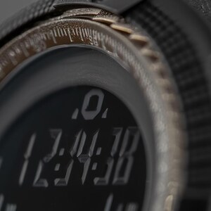 May include: A black digital watch with a textured bezel and a dark display screen showing the time 12:34:38 in a stylised font. The watch features a metallic bronze-toned inner ring with etched markings and a rugged, matte black casing.
