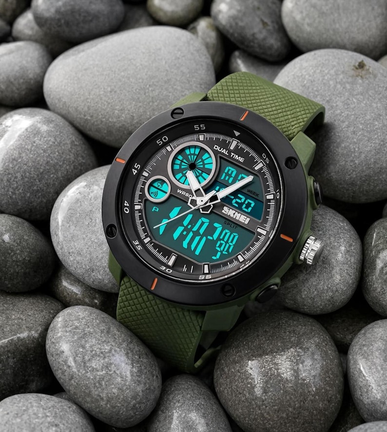 May include: A dual time wristwatch with an olive green textured silicone strap and a black circular bezel. The watch face features a combination of analog hands and a digital display with bright teal accents, displaying the brand name SKMEI.