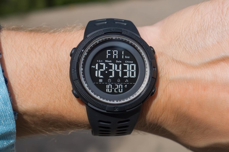 May include: A black digital sports watch with a round case and a textured silicone strap. The screen displays the time 12:34:38, the date 10-20, and the day FRI. The bezel features compass markings, and the interface includes icons for alarm and chime.