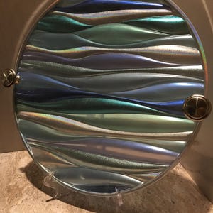 May include: A circular serving tray featuring a textured, wavy surface with a metallic finish in shades of blue, teal, and silver. The tray has a smooth, reflective border and two circular, metallic handles attached to the sides for easy carrying.
