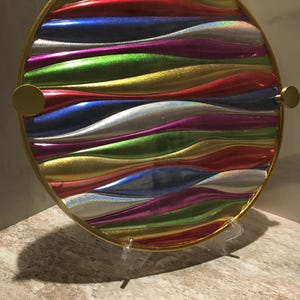 May include: A circular serving tray featuring a vibrant, multi-colored wavy pattern in shades of red, blue, green, silver, and gold. The tray has a metallic gold rim and two small gold circular handles on the sides, creating a reflective surface.