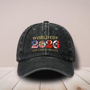 Könnte beinhalten: Eine schwarze Baseballkappe aus verwaschenem Denim mit dem goldenen Schriftzug WORLD CUP. Die Zahlen 2026 sind grafisch gestaltet und enthalten die Flaggen der USA, einen Fußball und Mexiko. Darunter steht der Text USA CANADA MEXICO.