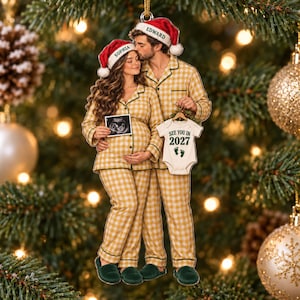 Peut inclure: Une décoration de Noël plate représentant un couple en pyjama à carreaux jaunes et blancs portant des bonnets de Père Noël étiquetés Sophia et Edward. La personne à gauche tient une échographie, tandis que celle de droite tient un body avec le texte See You In 2027 et des empreintes de pieds vertes.