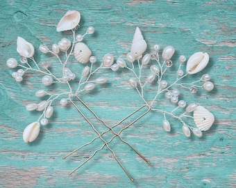 May include: Two gold hair pins with white seashells and pearls attached to them. The hair pins are arranged in a cross shape on a turquoise wooden background.