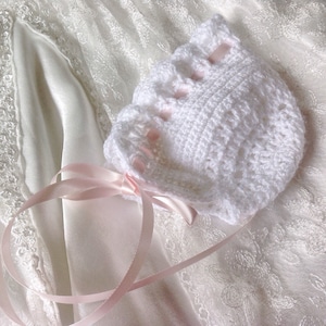 Puede incluir: Bonnet de bebé de ganchillo blanco con una cinta de raso rosa. El gorro tiene un borde festoneado y está decorado con lazos de raso rosa.