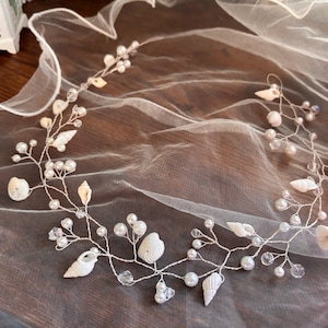 Puede incluir: Una delicada enredadera para el cabello de novia con conchas blancas, perlas de imitación y cuentas de cristal transparente. El alambre es plateado y el diseño está dispuesto en un patrón fluido y orgánico. El accesorio para el cabello está sobre una tela blanca transparente.