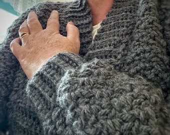 Könnte beinhalten: Ein grauer Strickcardigan mit einem strukturierten Muster. Der Cardigan hat einen V-Ausschnitt und lange Ärmel.