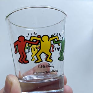 Può includere: Un bicchiere da shot in vetro trasparente con una grafica colorata di Keith Haring raffigurante figure stilizzate in rosso, giallo e verde che si tengono per mano. Il vetro riporta la firma stampata Keith Haring e il testo Estate of Keith Haring.
