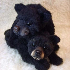 以下が含まれることがあります： 2匹の黒いぬいぐるみクマ。1匹がもう1匹の上に座っている。上のクマは頭を横に傾け、目を閉じている。下のクマは目を大きく開けてカメラを見ている。