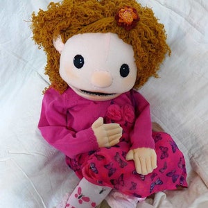 May include: A handmade doll with a plush face, curly orange hair, and a pink outfit. The doll wears a pink sweater, a dress with butterfly patterns, and matching socks and shoes. The doll's features include large black eyes and a small nose.