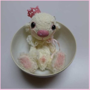 以下が含まれることがあります： 白いセラミックボウルの中に、ピンクのアクセントが付いた小さな白いぬいぐるみが座っています。おもちゃは頭に王冠をかぶっており、大きく黒い目を持っています。ピンクと白のフリル付きの襟をしています。