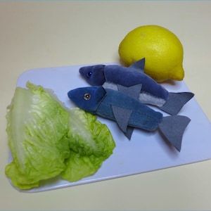 以下が含まれることがあります： 白いまな板の上に、白いアクセントのある青いぬいぐるみ製の魚が2匹と、緑のレタスと黄色のレモンが置かれています。