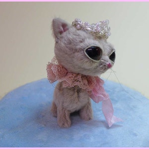 以下が含まれることがあります： ピンクのレースの襟と銀と白のビーズの冠をかぶった灰色のぬいぐるみ猫。猫は大きな黒い目とピンクの鼻をしています。猫は薄い青色のふわふわした表面に座っています。