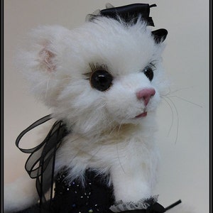 以下が含まれることがあります： 黒いビーズと黒いリボンが付いた黒いドレスを着た白いぬいぐるみ猫。猫は黒い目とまつげ、そしてピンクの鼻をしています。