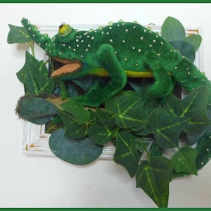 以下が含まれることがあります： 白い額縁の中に、緑色のカメレオンのぬいぐるみが緑の葉っぱの上に置かれています。カメレオンのぬいぐるみは金色のキラキラした装飾が施されています。