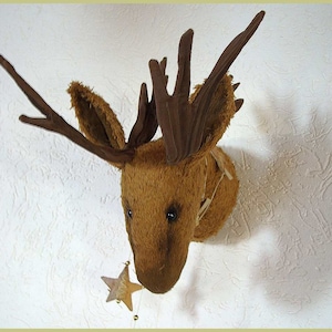 以下が含まれることがあります： 白い壁に、大きな角のついた茶色のぬいぐるみ製のヘラジカの頭が取り付けられています。ヘラジカの頭の真下に、小さな金の星がぶら下がっています。