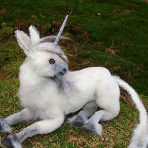 以下が含まれることがあります： 銀色の角と長い白い尻尾を持つ白いぬいぐるみユニコーン。ユニコーンは緑の苔のベッドの上に横たわっています。