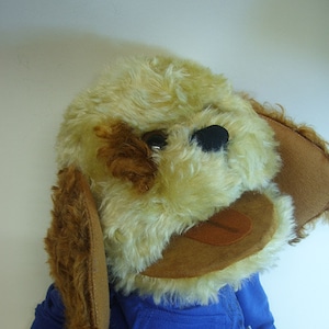 May include: A plush dog puppet with a tan and brown fur face, brown ears, and a blue hoodie. The puppet has a smiling expression with a brown tongue. The background is plain white.