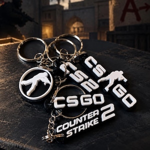Puede incluir: Un conjunto de llaveros en blanco y negro con el texto CSGO, Counter Strike 2 y la silueta de un soldado apuntando con un rifle. Cada llavero está unido a una anilla de metal plateado, mostrando letras blancas sobre una base negra.