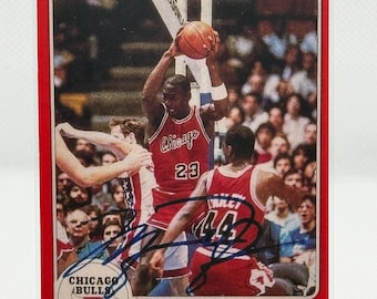 May include: A 1985 Star basketball trading card featuring Michael Jordan in a red Chicago Bulls uniform, number 23, holding a basketball. The card includes a black ink autograph, the text STAR '85, Chicago Bulls, and Michael Jordan Guard.