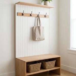 Peut inclure: Un banc d'entrée en bois clair avec un panneau arrière en lambris blanc, une étagère supérieure et quatre crochets en métal noir. Un sac fourre-tout beige est suspendu à un crochet. La base comporte deux compartiments de rangement ouverts.