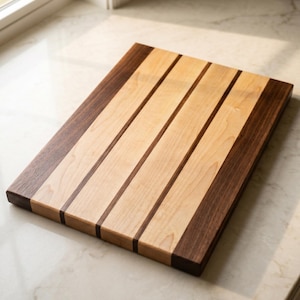 May include: A rectangular wooden cutting board featuring a striped pattern of alternating light maple and dark walnut wood planks. The board has a smooth, flat surface with visible wood grain and clean, straight edges, showcasing a natural contrast.