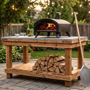 May include: A black outdoor pizza oven with a burning fire inside sits on a rectangular wooden table with a stone top. The table features a lower shelf stacked with firewood and a long-handled metal pizza peel leaning against the side.