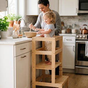 May include: A light-colored wooden learning tower with a rectangular frame, a raised standing platform, and a lower step. The structure features vertical support posts, horizontal safety rails, and a sturdy base designed to provide elevation.
