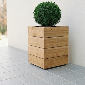 Könnte beinhalten: Ein quadratischer Pflanzkübel aus Holz, der aus vier horizontalen Planken aus hellbraunem Naturholz gefertigt ist. Der Kübel hat ein klares, geometrisches Design und enthält einen dichten, kugelförmigen grünen Strauch mit kleinen, abgerundeten Blättern.