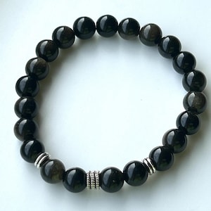 May include: A stretch bracelet composed of smooth, round, black obsidian beads with a subtle sheen. The design features two silver-toned metal spacer beads with a ribbed texture, positioned symmetrically to accent the dark stone beads.