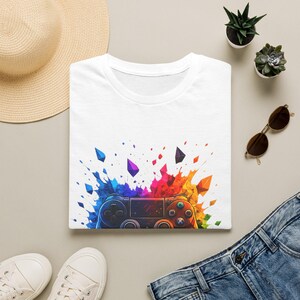 Könnte beinhalten: Ein weißes T-Shirt mit einem Grafikdruck eines Videospiel-Controllers, der von einem lebendigen, mehrfarbigen Farbspritzer-Effekt in Blau-, Rot-, Gelb- und Orangetönen umgeben ist, wobei geometrische Formen über dem Controller schweben.
