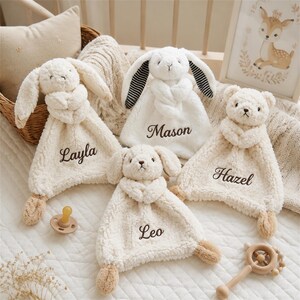 May include: Four cream-colored plush security blankets featuring animal heads and embroidered names. The blankets are labeled Layla, Mason, Hazel, and Leo. Each soft, textured blanket includes a stuffed animal top and small paws at the corners.