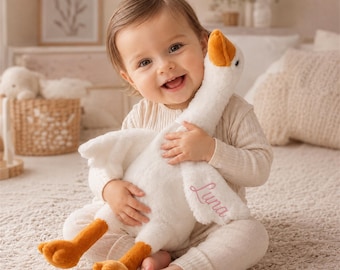 May include: A plush white goose toy with an orange beak and orange feet. The wing of the goose features the name Luna embroidered in pink script. The toy has a soft, fuzzy texture and is designed with a long neck and outstretched wings.