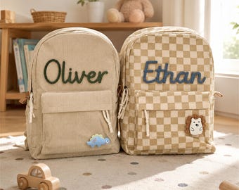 May include: Two corduroy backpacks, one solid beige with a green embroidered name Oliver and a blue dinosaur patch, and one beige checkered with a blue embroidered name Ethan and a hedgehog patch. Both feature front pockets and white rope zippers.