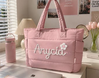 May include: A dusty pink quilted tote bag featuring two top handles and side pockets. The front of the bag displays the white embroidered name Anycia in a cursive script, accompanied by a small white and yellow crocheted flower appliqué.