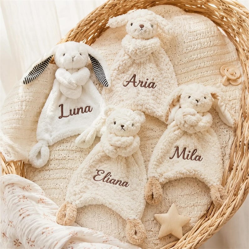 May include: Four plush security blankets featuring stuffed animal heads and embroidered names. One white rabbit with striped ears labeled Luna, one cream lamb labeled Aria, one cream bear labeled Eliana, and one cream rabbit labeled Mila.
