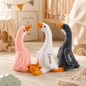 Puede incluir: Tres gansos de peluche con pelaje suave en rosa, blanco y gris oscuro. Cada ganso tiene un pico y patas de color naranja, con un nombre bordado en el ala: Quinn en el ganso rosa, Luna en el blanco y Sophie en el ganso gris oscuro.