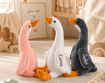 May include: Three plush goose toys with soft fur in pink, white, and dark gray. Each goose features an orange beak and feet, with a name embroidered on the wing: Quinn on the pink goose, Luna on the white goose, and Sophie on the dark gray goose.