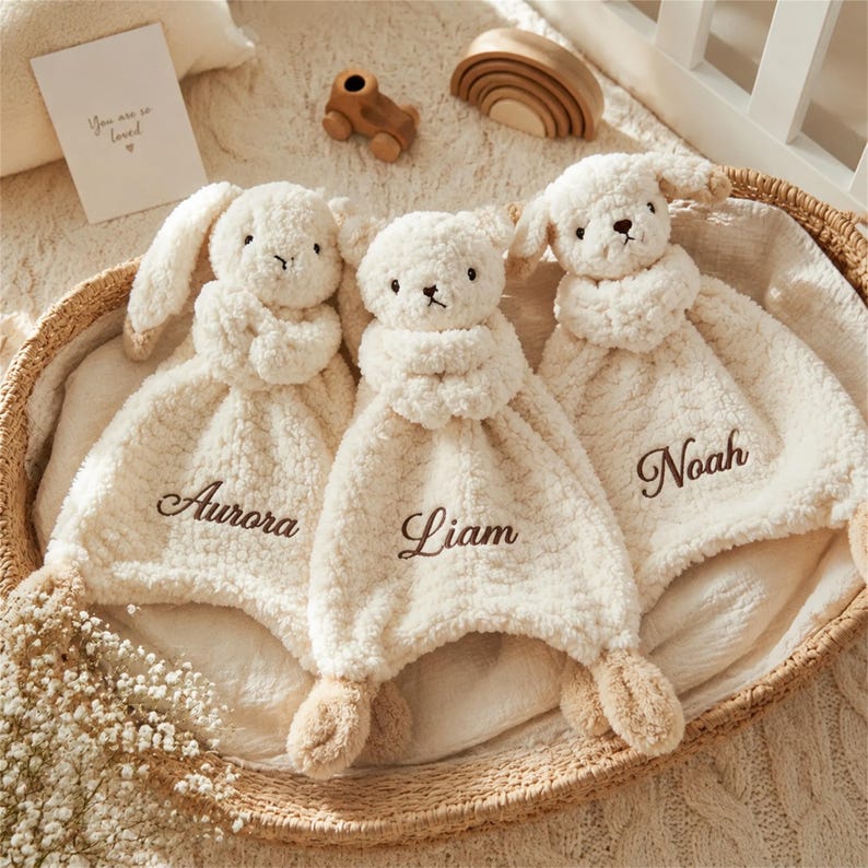 May include: Three cream-colored plush security blankets featuring animal heads and embroidered names. The left blanket is embroidered with Aurora, the center with Liam, and the right with Noah. Each soft, textured blanket has a small animal head.
