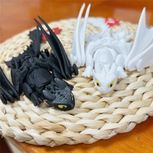 Peut inclure: Deux figurines de dragon articulées, une noir mat et une blanc mat, dotées de grandes ailes, d'écailles texturées et d'yeux jaune vif. Les figurines sont placées côte à côte, mettant en valeur leurs membres et têtes détaillés et mobiles.