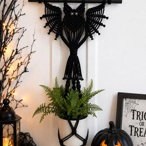 May include: A black macrame wall hanging shaped like a bat with two small silver eyes, suspended from a black wooden rod. The macrame cord extends downward to form a plant hanger holding a white ceramic pot containing a green fern plant.