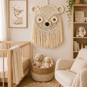 May include: A cream-colored macrame wall hanging shaped like a bear face, featuring intricate knotted patterns, circular ears, two wooden bead eyes, and a dark nose. The piece includes long, flowing fringe at the bottom and is suspended by a cord.