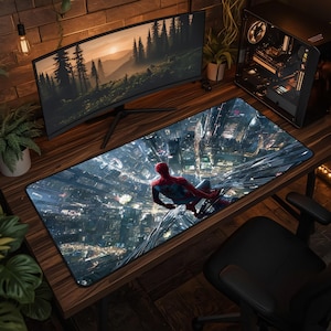 May include: A rectangular desk mat featuring a high-resolution graphic of the superhero Spider-Man perched on a skyscraper overlooking a sprawling city at night. The mat has a black stitched border and rests on a dark wood grain desk surface.