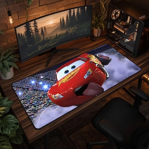 May include: A rectangular desk mat featuring a large, red, anthropomorphic race car with blue eyes, a wide mouth, and the number 95 on its hood. The car is depicted on a racetrack with a crowd of spectators in the background, rendered in vibrant colors.