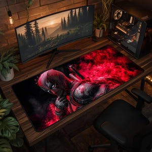 May include: A rectangular desk mat featuring a graphic illustration of the character Deadpool in a red and black suit, surrounded by vibrant red smoke effects. The mat has a black border and is positioned on a wooden desk surface.