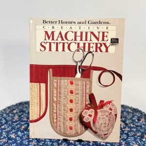 May include: A hardcover book titled Better Homes and Gardens Creative Machine Stitchery. The cover features a beige pocket with red trim, silver scissors, and a heart-shaped lace ornament with a red ribbon against a light background.