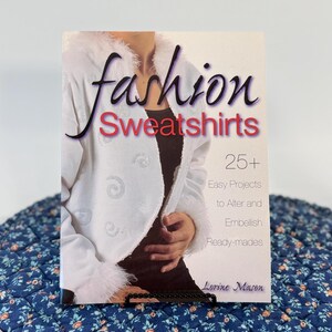 May include: A softcover book titled Fashion Sweatshirts by Lorine Mason, featuring 25+ Easy Projects to Alter and Embellish Ready-mades. The cover displays a white sweatshirt with a fuzzy collar and cuffs, decorated with subtle silver swirl patterns.