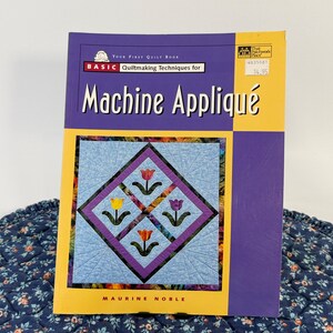May include: A softcover instructional book titled Your First Quilt Book Basic Quiltmaking Techniques for Machine Appliqué by Maurine Noble. The cover features a light blue quilt block with four colorful tulip designs and purple borders.