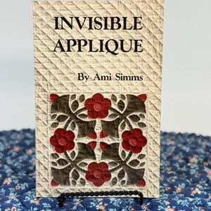 May include: A book titled Invisible Applique by Ami Simms with a cream-colored quilted cover featuring a central square block of dark brown and red floral applique patterns. The book is displayed on a black wire stand against a white background.