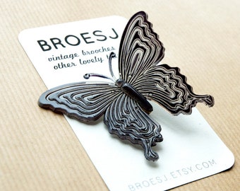 May include: A dark gray butterfly brooch with a layered design, resting on a white card. The card features the text "BROESJ vintage brooches other lovely things" and the website address. The brooch is a vintage accessory.